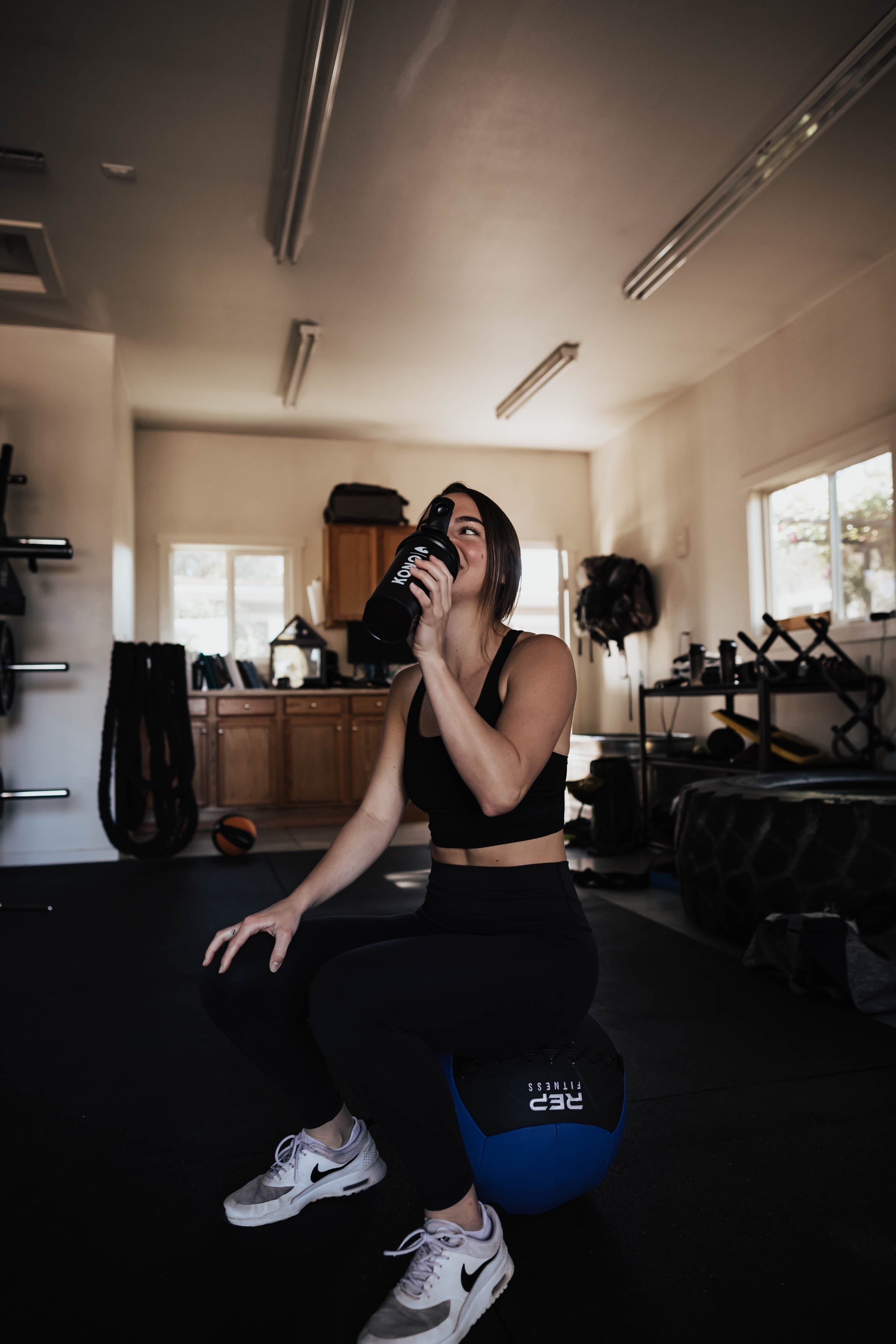 gym lifestyle photo—female athlete hydrating with KONO shaker while resting on a medicine ball