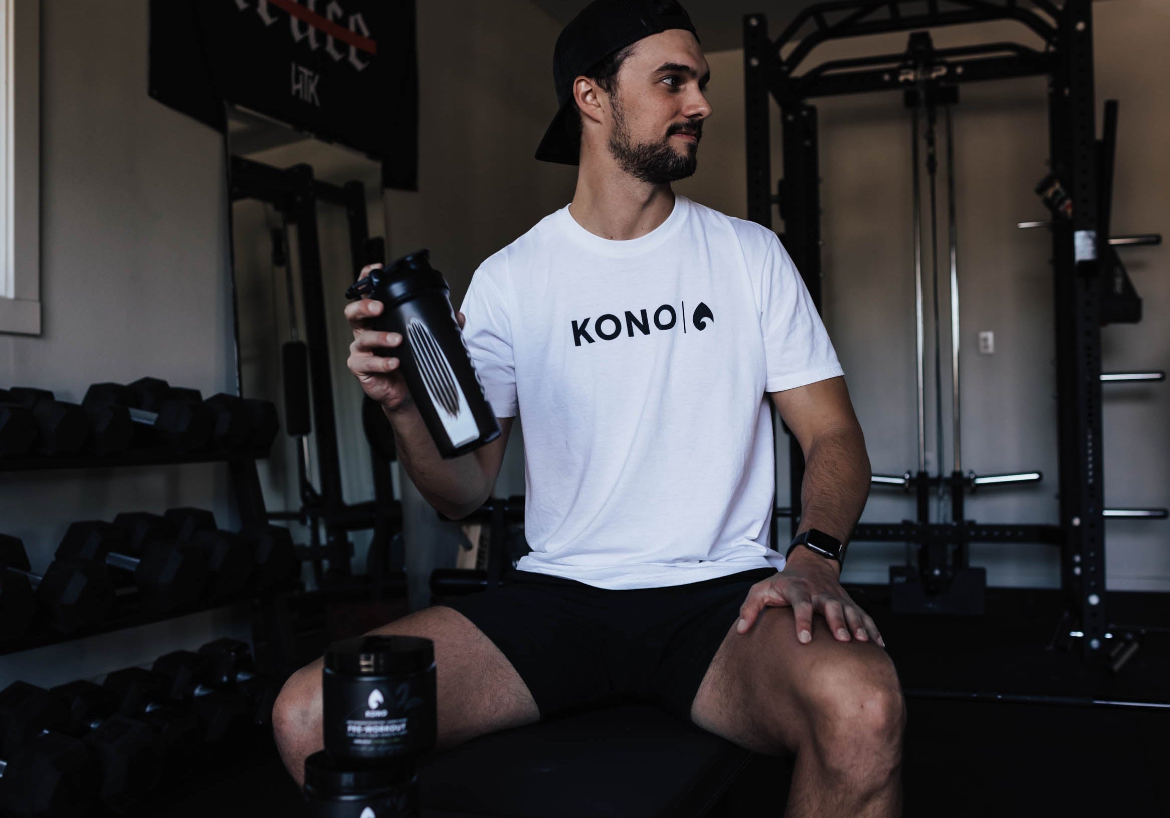 athlete sitting on a weight bench in a home gym holding a black KONO shaker, pre-workout tubs on the bench, racks and dumbbells behind