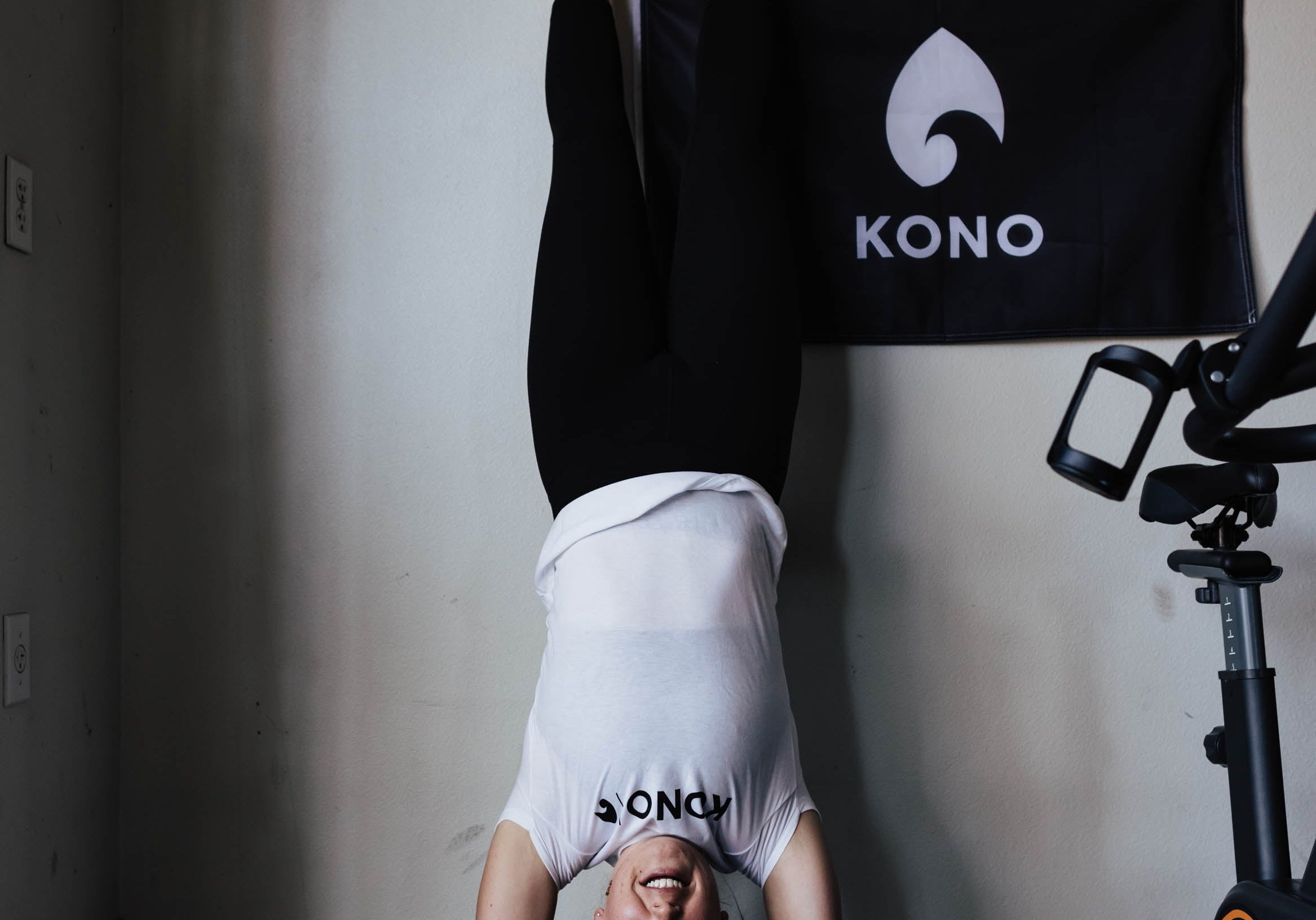 woman in a KONO shirt performing a wall handstand on a blue pad in a garage gym with REP Garage Gym and KONO banners