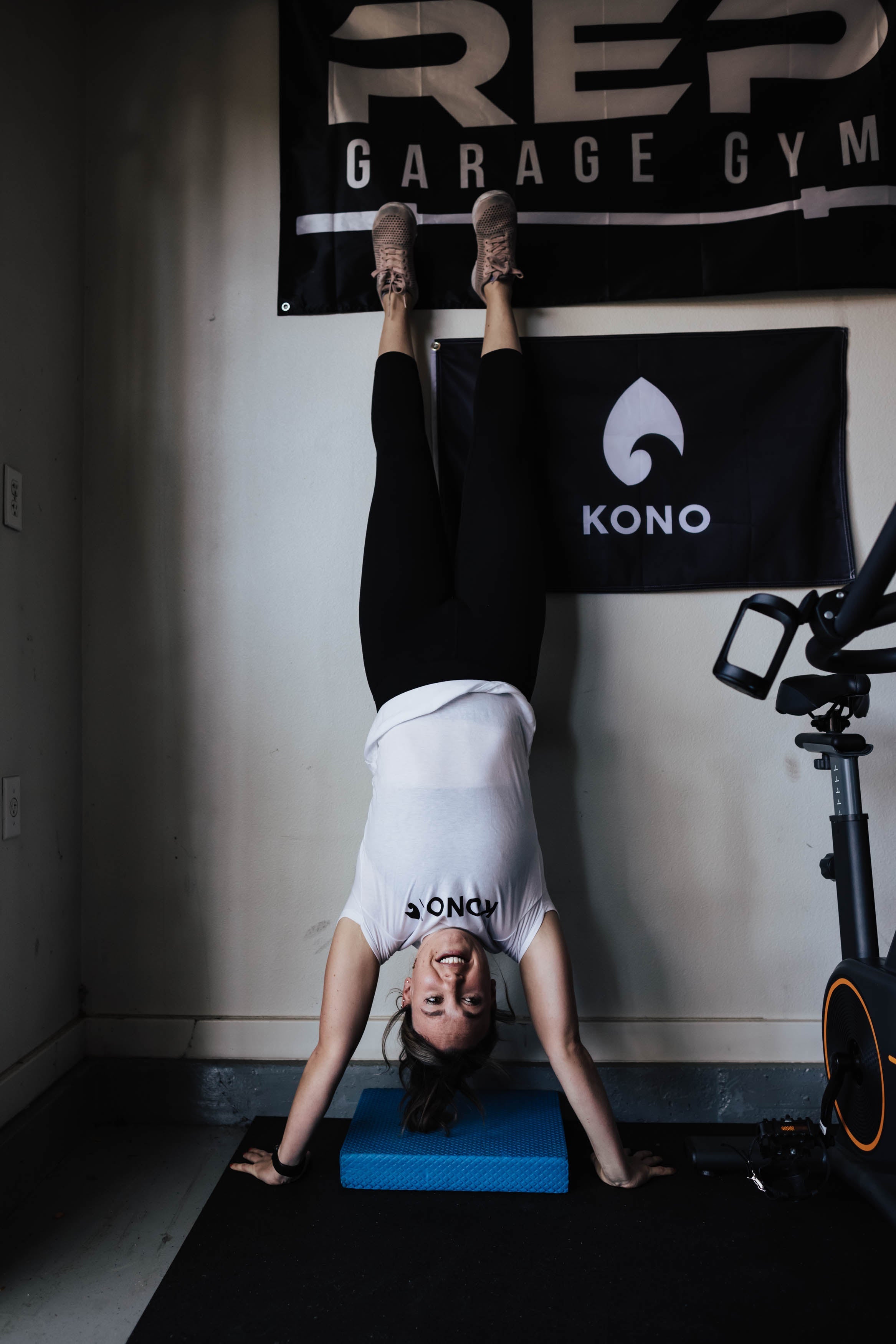woman in a KONO shirt performing a wall handstand on a blue pad in a garage gym with REP Garage Gym and KONO banners