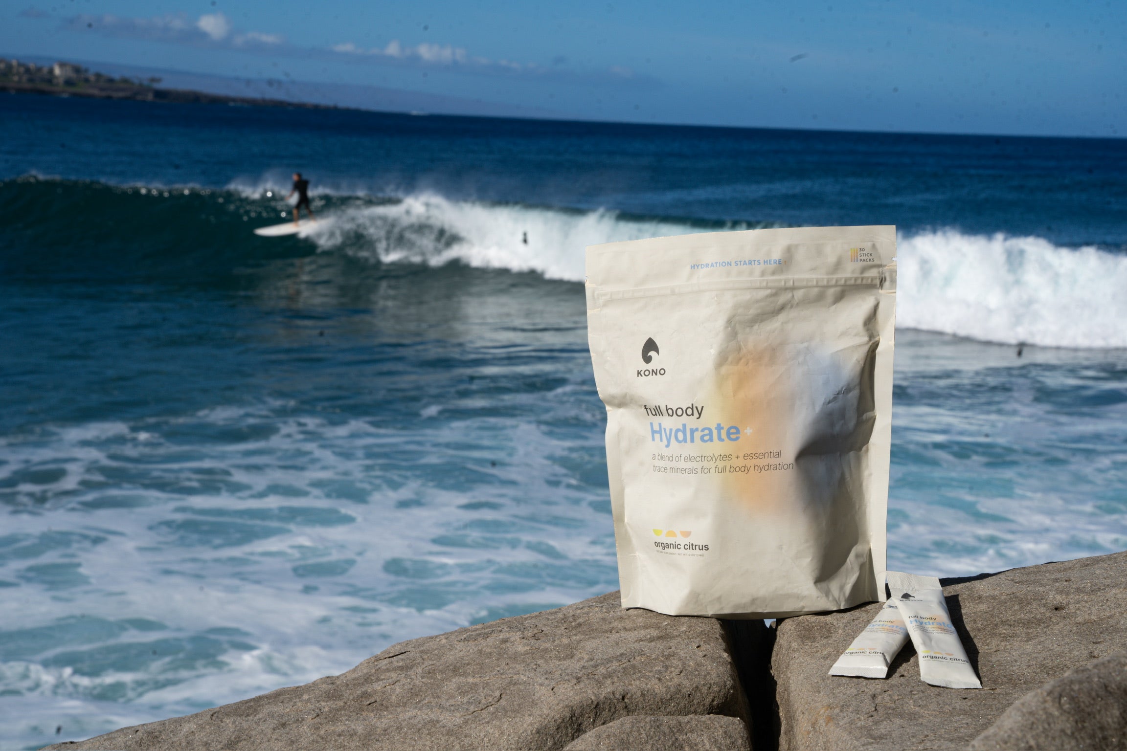 KONO Hydrate+ electrolyte powder pouch and stick packs on coastal rocks with ocean waves and a surfer in the background