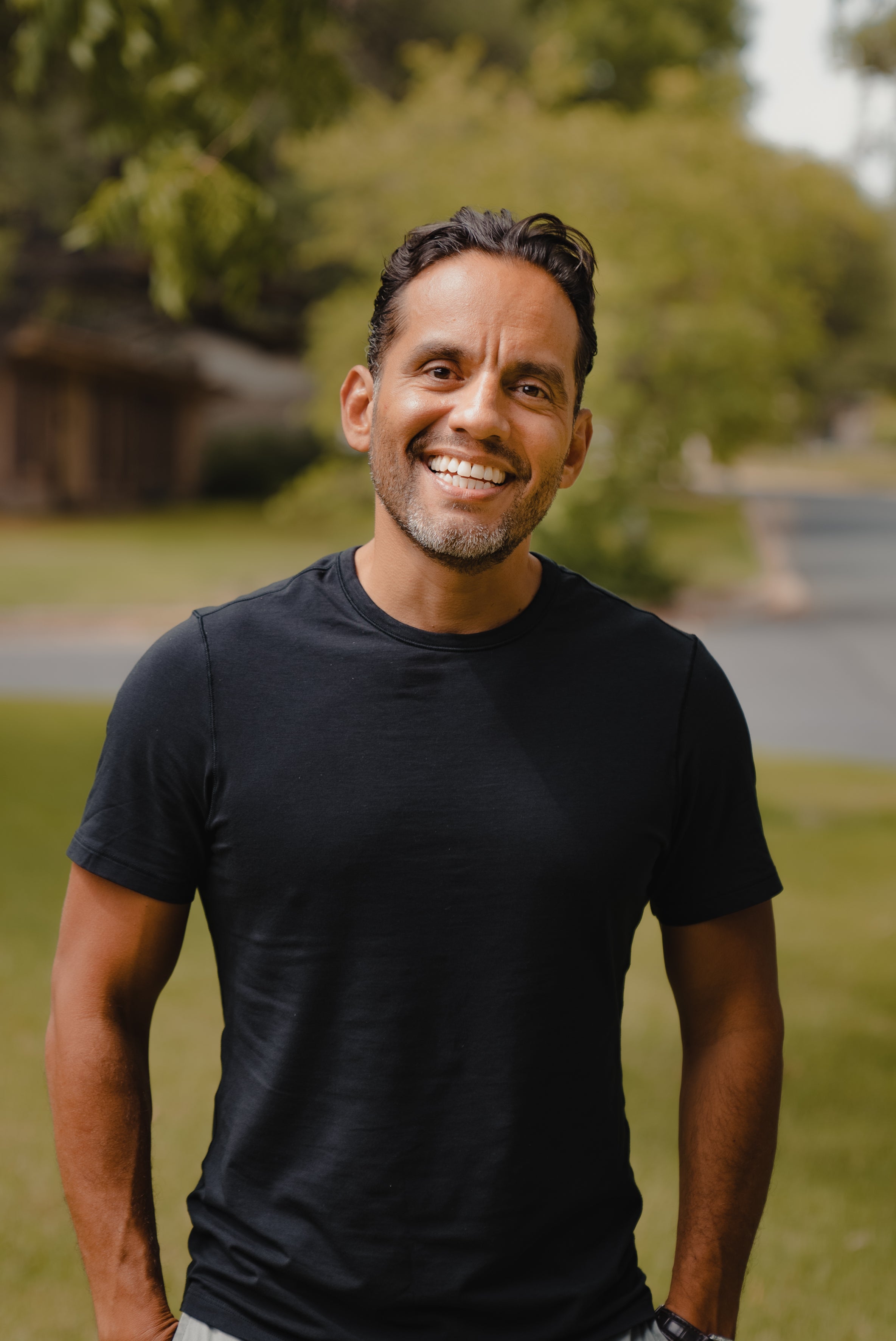 casual outdoor headshot of a man standing in a park with trees and path blurred behind