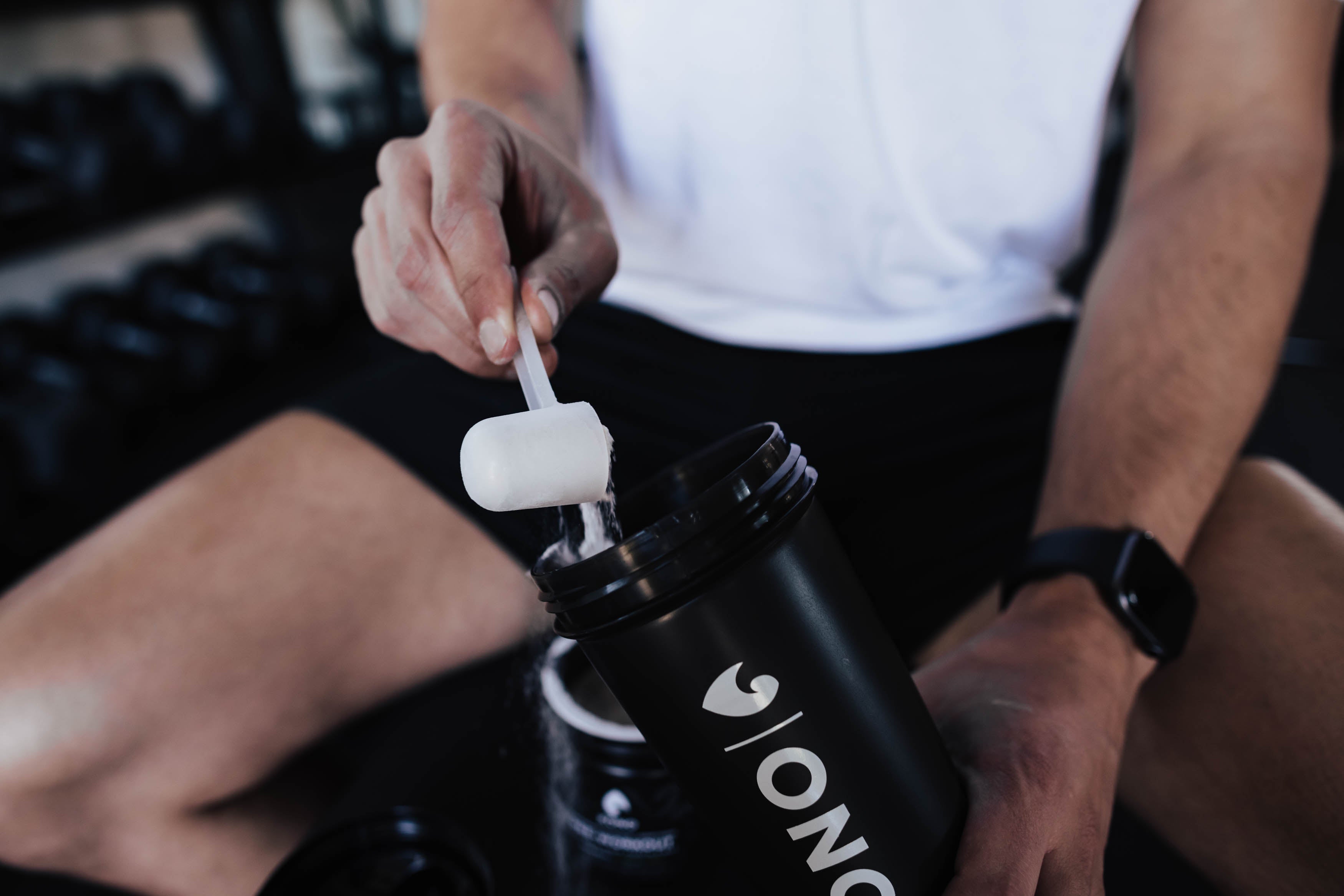 athlete mixing KONO pre-workout powder into a black shaker bottle at the gym, close-up hand with scoop