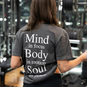 Woman in gym wearing charcoal KONO tee showing large motivational back print—‘Mind in focus, Body in motion, Soul in peace.’