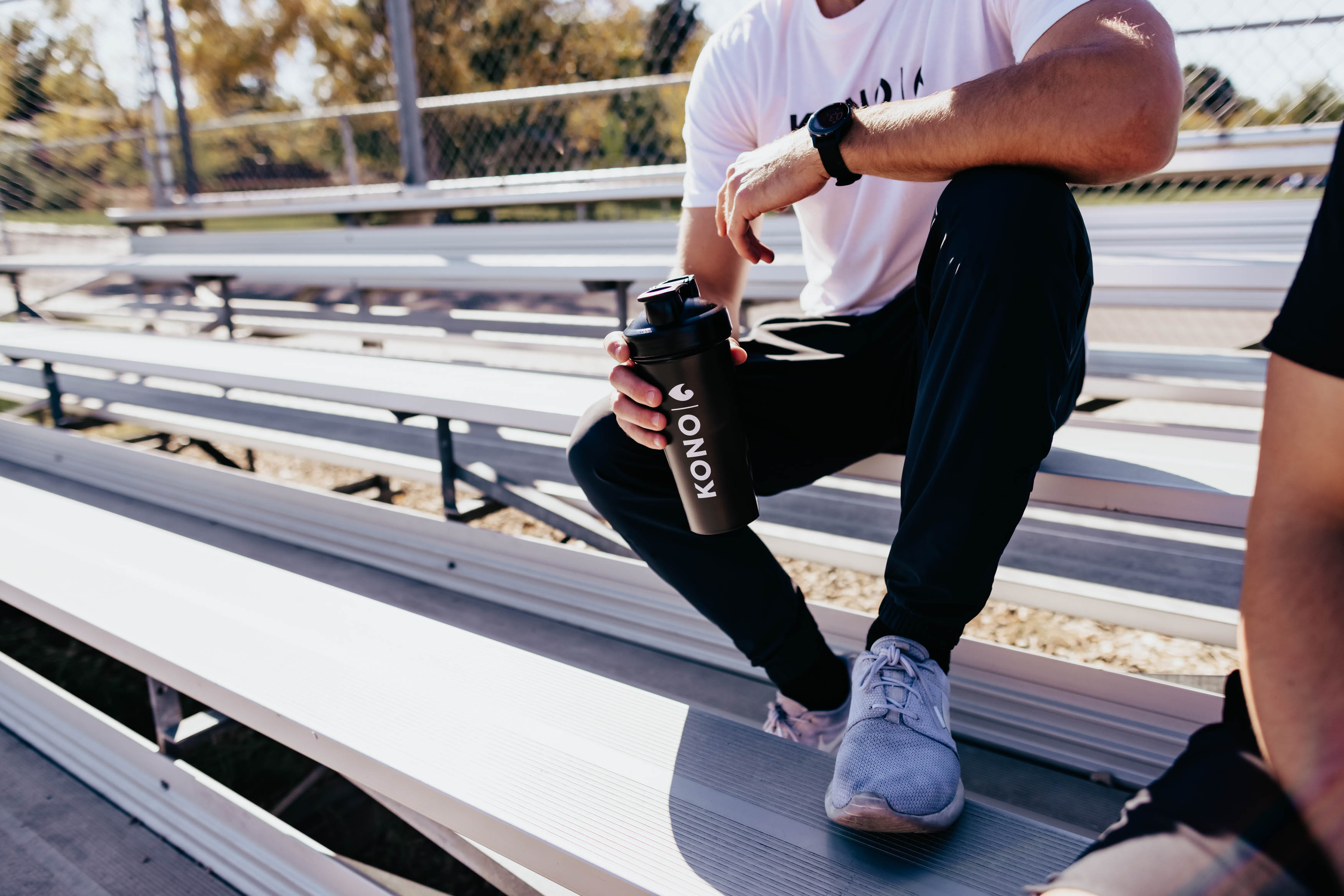 fitness lifestyle shot—person in KONO tee and sneakers resting on bleachers with KONO shaker cup