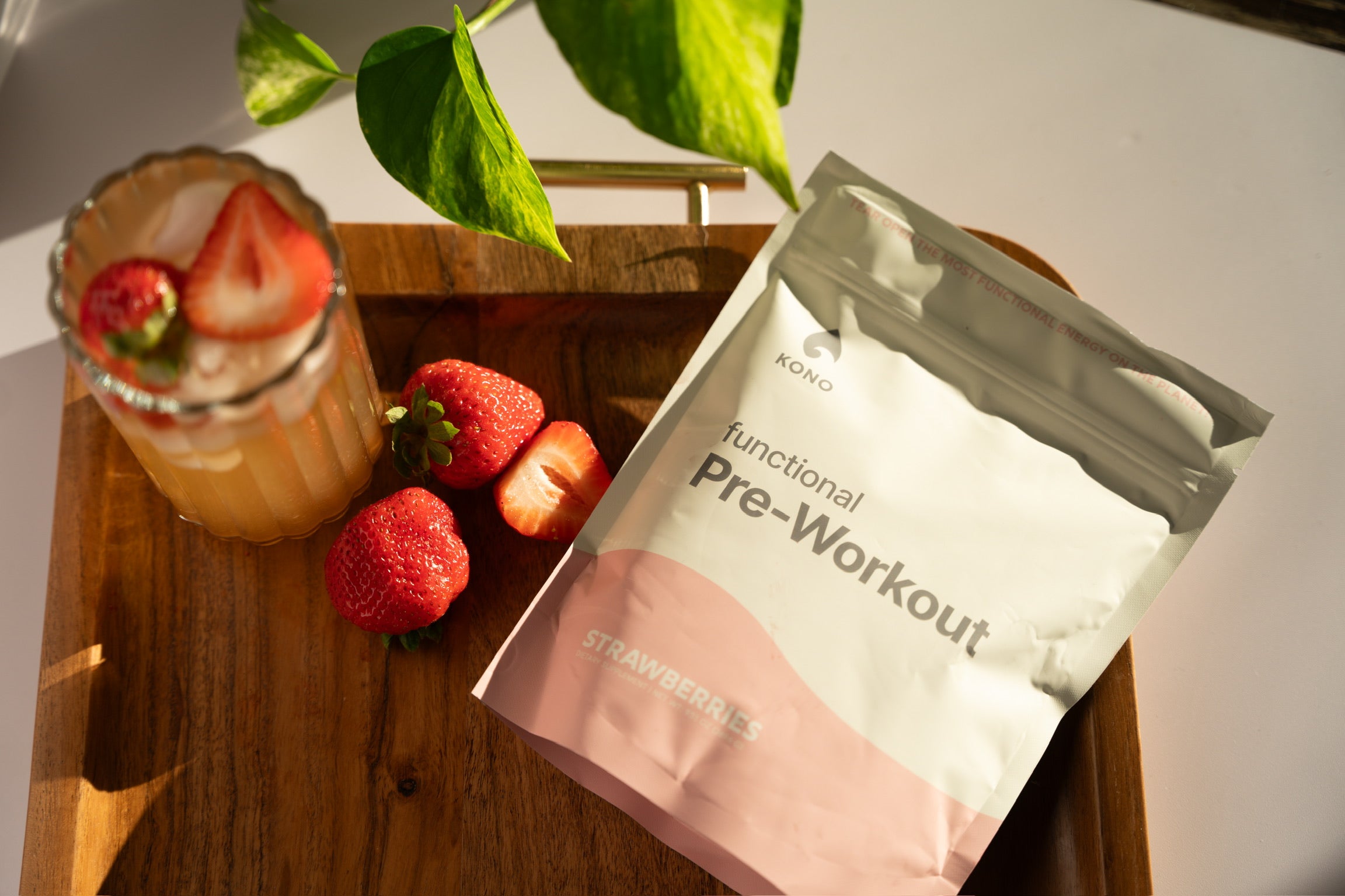 KONO functional pre-workout (strawberries flavor) pouch on a wooden tray beside fresh strawberries and a strawberry-garnished iced drink in warm sunlight
