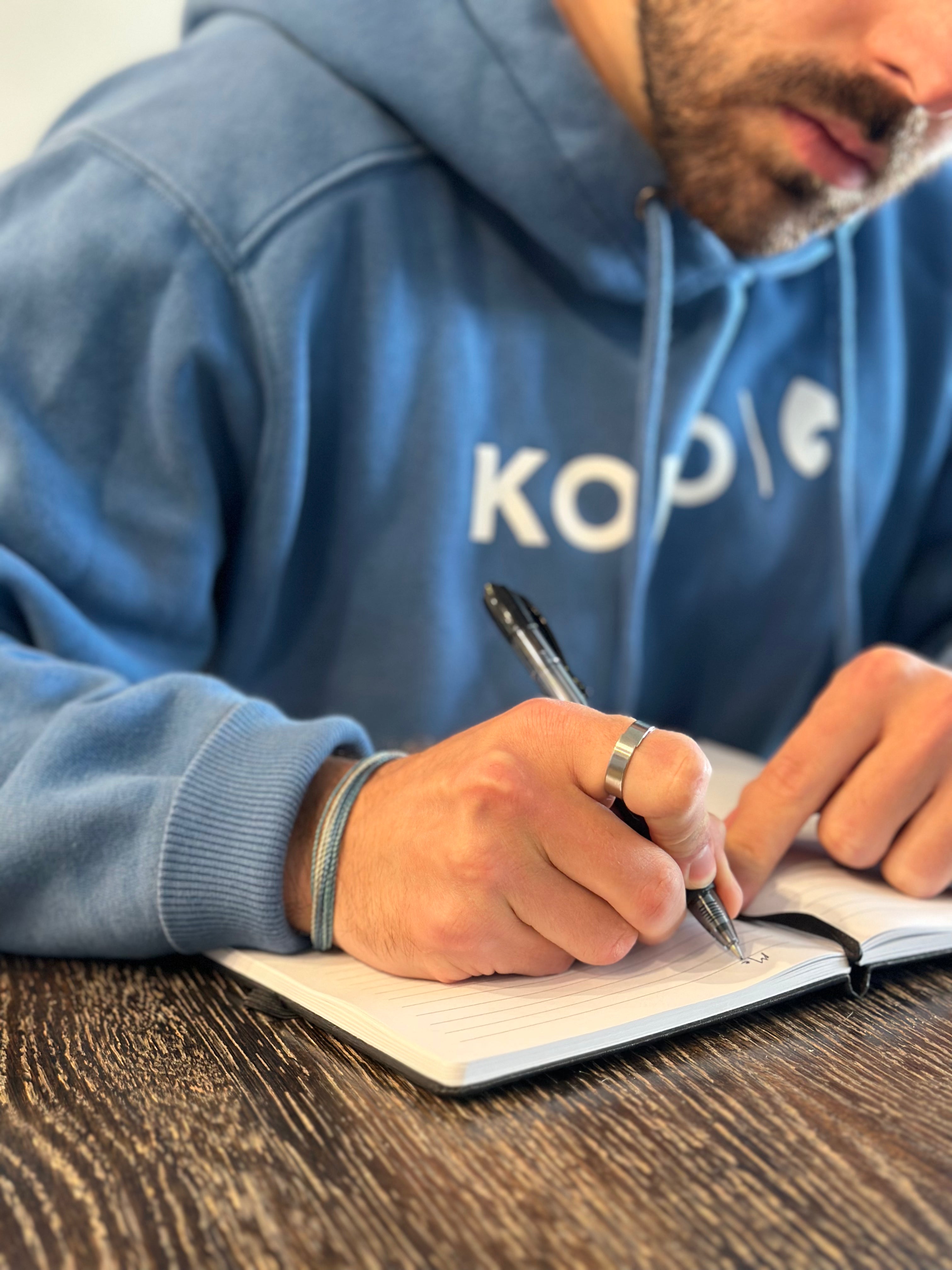 Man in blue KONO hoodie writing in a lined notebook at a wooden table, close-up of hands, pen, and chest logo.