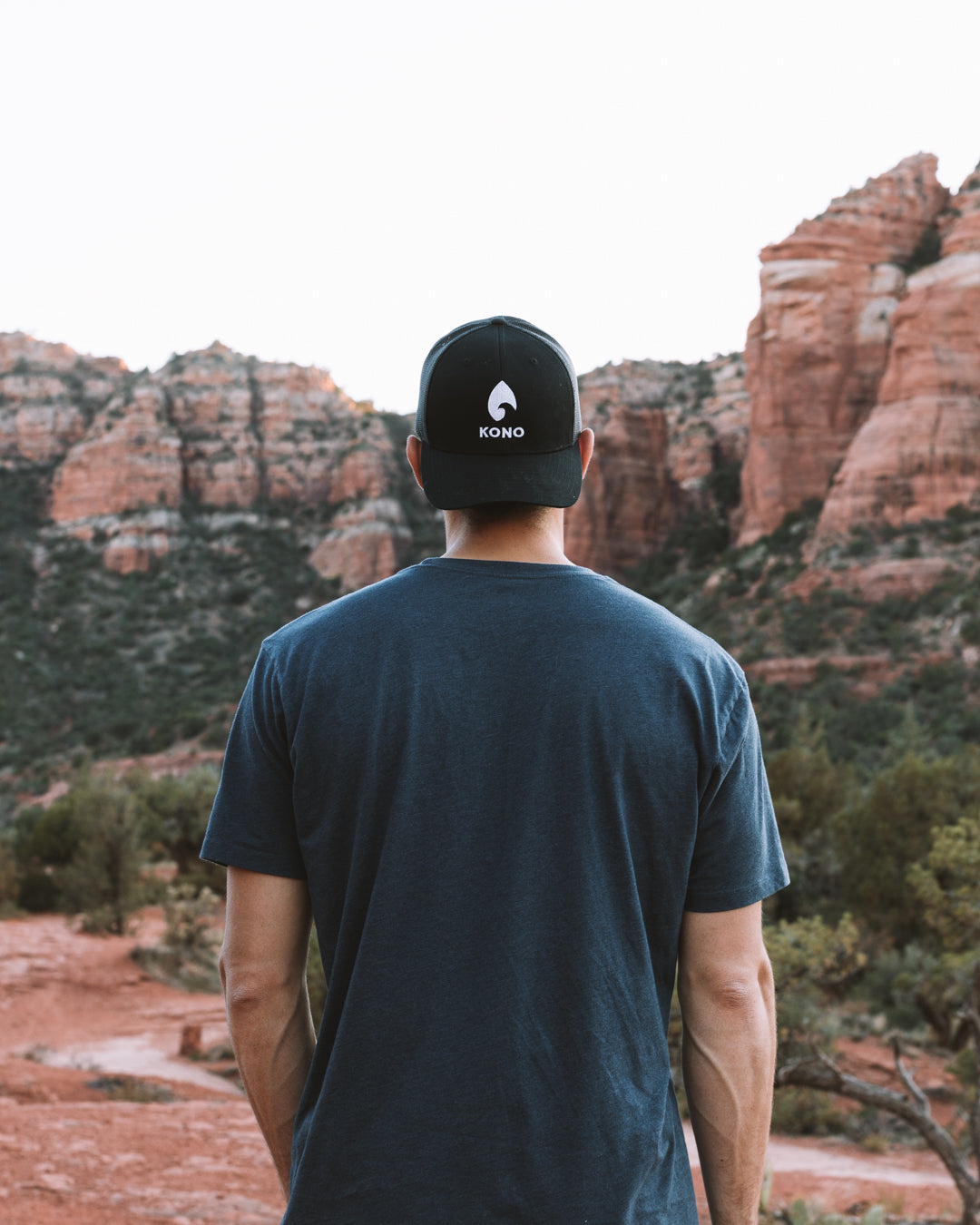 Man from behind in a black KONO logo trucker hat standing in a red-rock canyon—adventure/outdoor lifestyle.