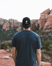 Man from behind in a black KONO logo trucker hat standing in a red-rock canyon—adventure/outdoor lifestyle.