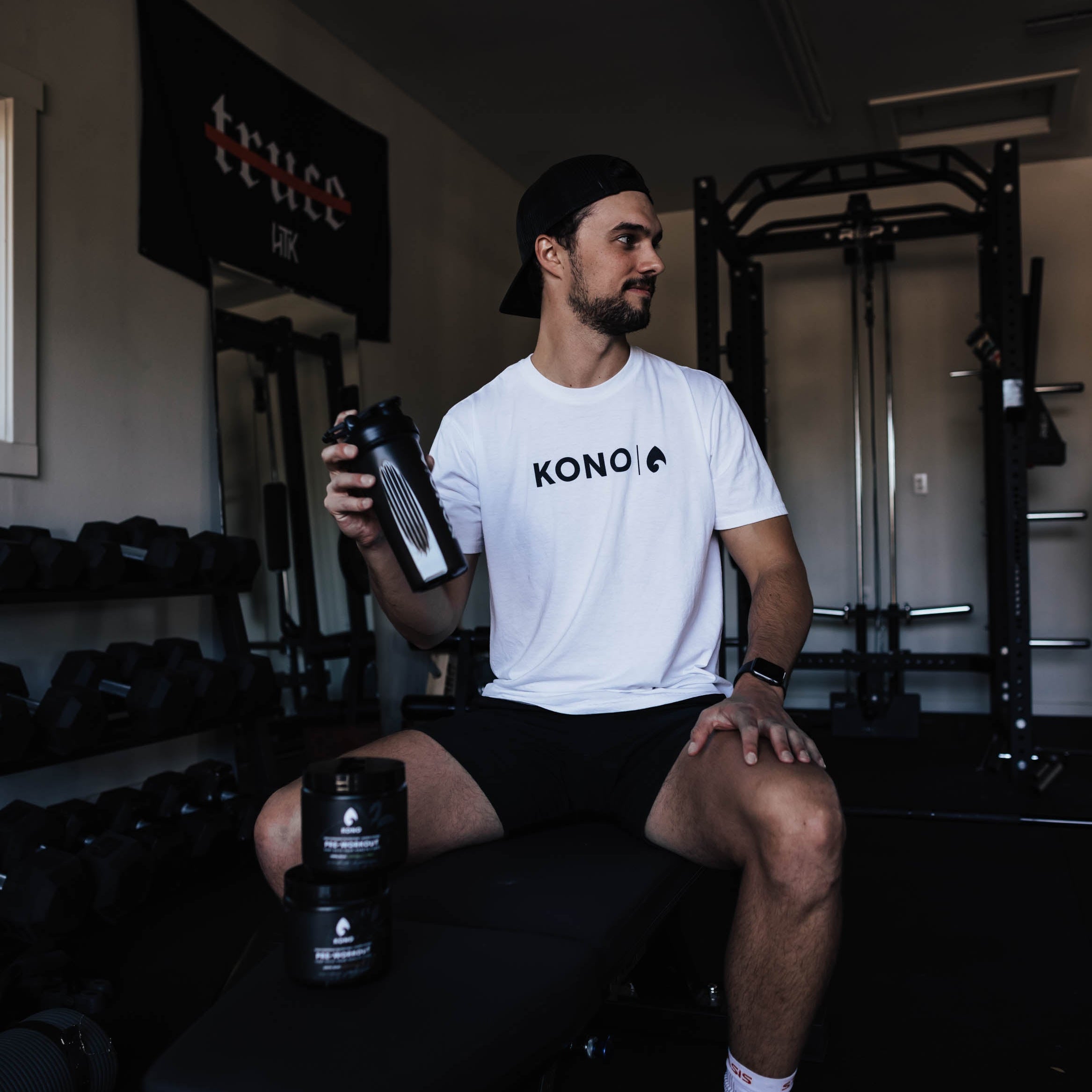 athlete sitting on a weight bench in a home gym holding a black KONO shaker, pre-workout tubs on the bench, racks and dumbbells behind