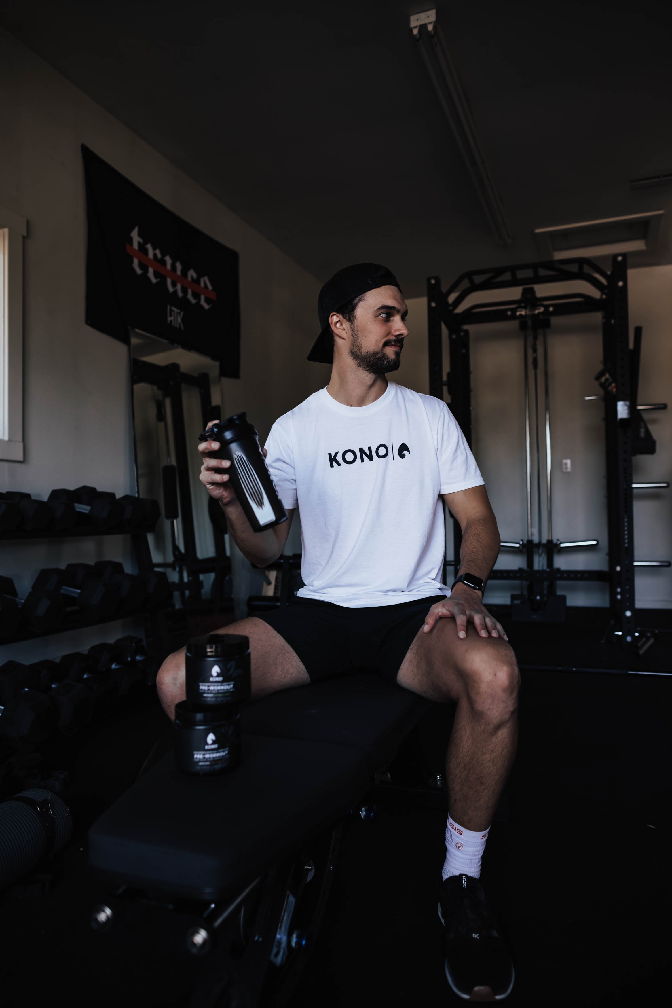 athlete sitting on a weight bench in a home gym holding a black KONO shaker, pre-workout tubs on the bench, racks and dumbbells behind