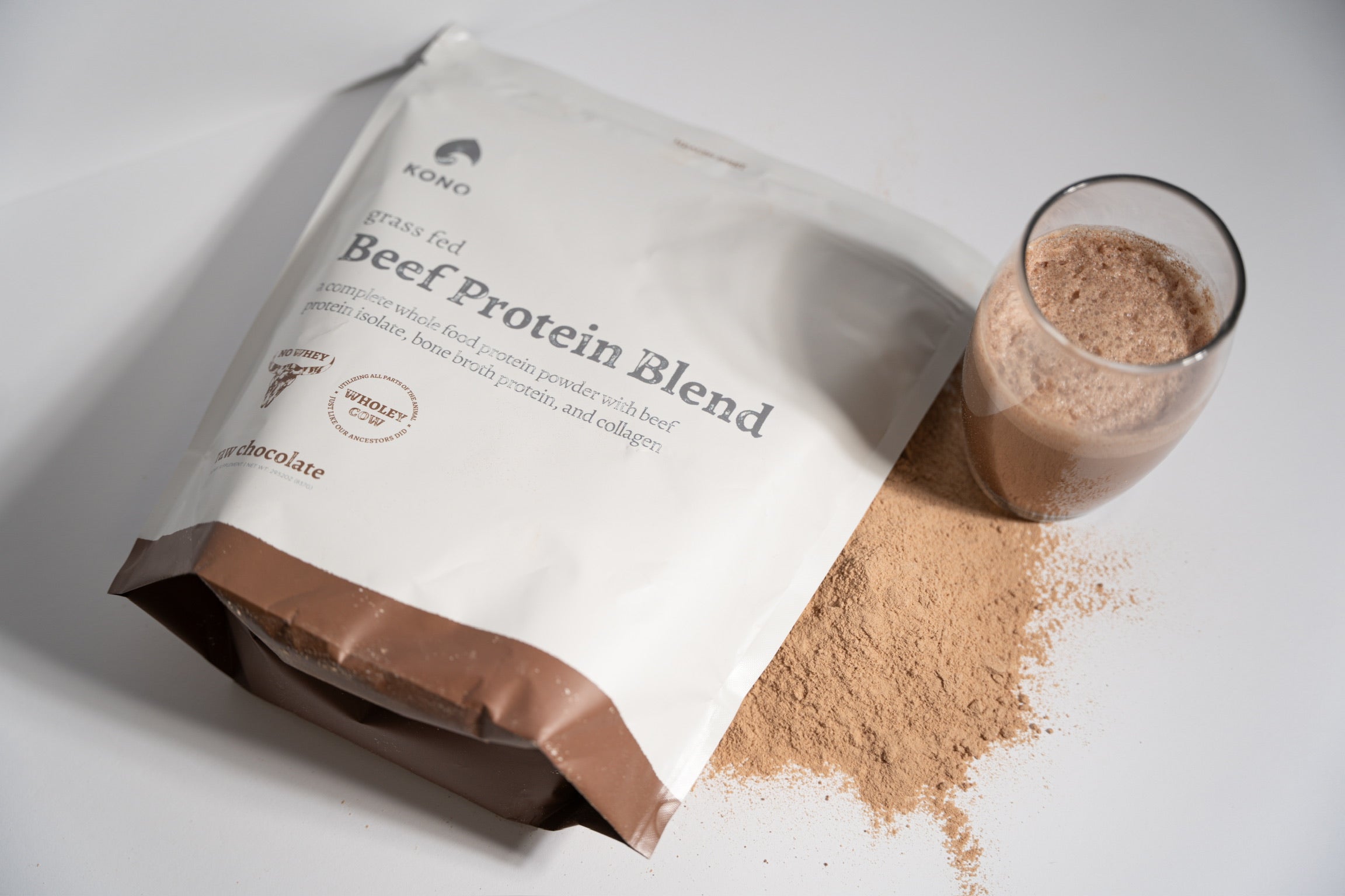 KONO grass-fed Beef Protein Blend (chocolate) pouch with spilled powder and a glass of mixed shake on a white background