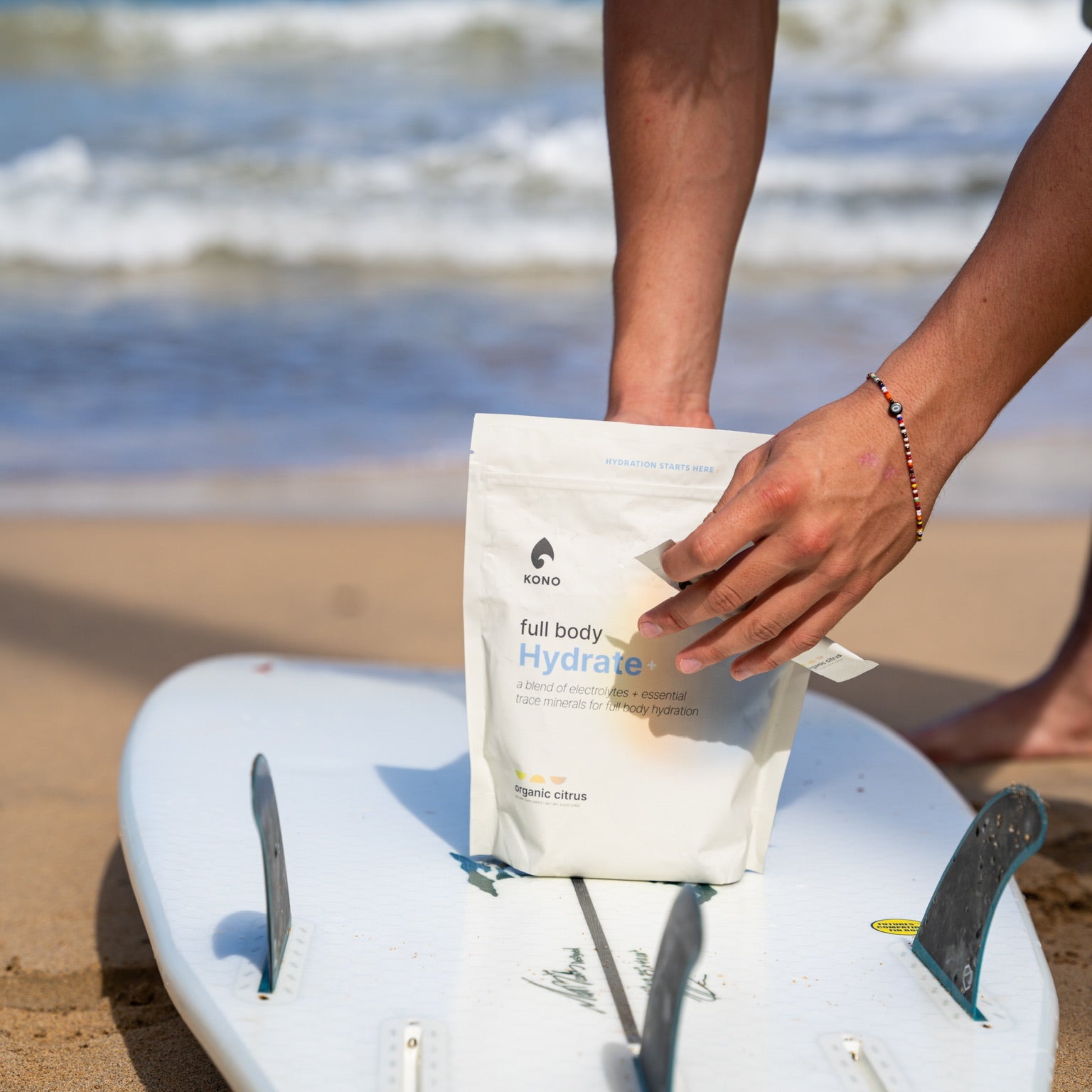 hand pulling a stick pack from a KONO Hydrate+ pouch placed on a white surfboard at the shoreline, ocean waves in the background