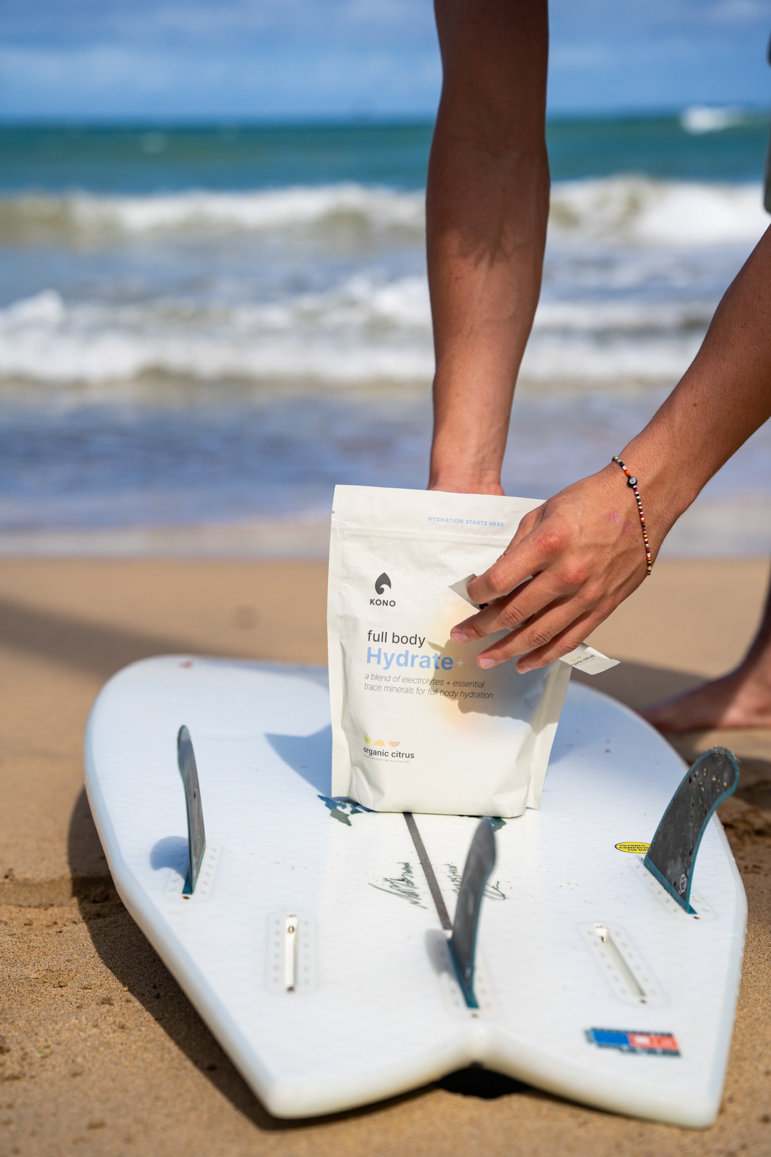 hand pulling a stick pack from a KONO Hydrate+ pouch placed on a white surfboard at the shoreline, ocean waves in the background