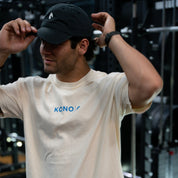 Model wearing a black KONO logo dad hat and cream KONO tee in a gym—fitness lifestyle apparel photo.