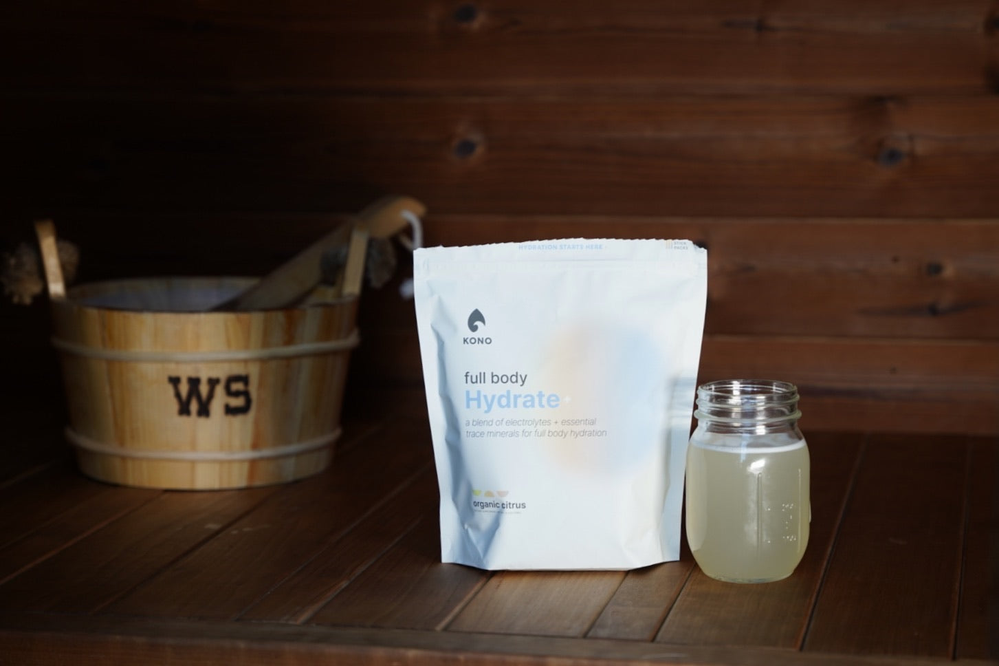 sauna recovery scene with KONO Hydrate+ packaging and a glass jar of citrus electrolyte mix on wooden slats