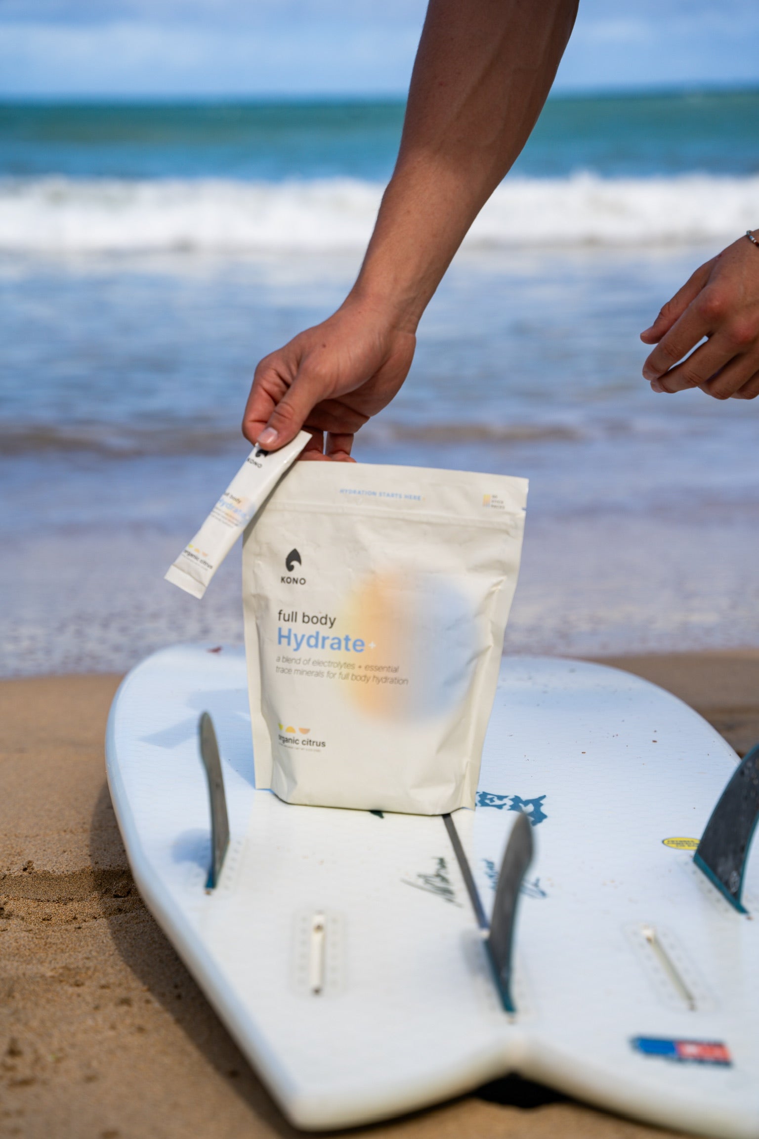 Kono Hydrate pouch placed on a surfboard at the beach with ocean waves in the background.