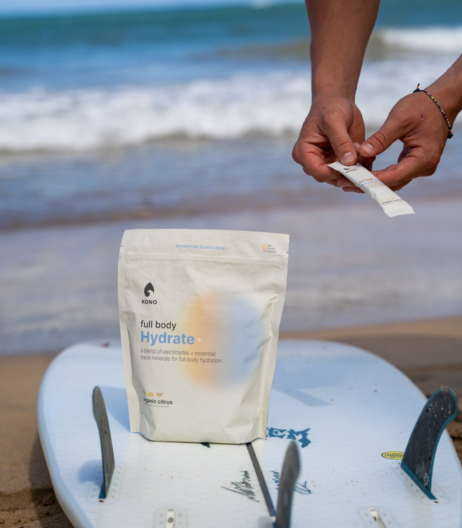 beach lifestyle shot—KONO Hydrate+ organic citrus pouch on surfboard with waves in background