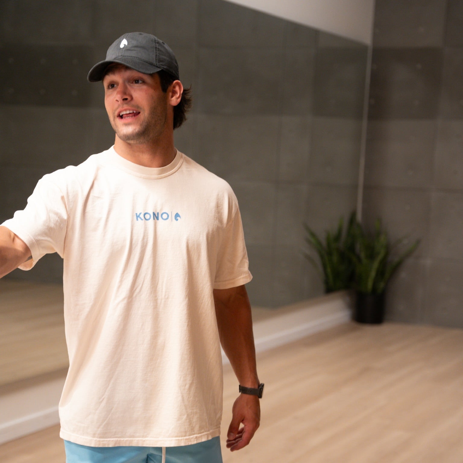 Man in cream KONO logo t-shirt and cap in studio gym—athleisure top, relaxed fit.