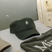 KONO forest-green dad hat with embroidered logo on table beside KONO whey protein pouches and apparel—lifestyle scene.