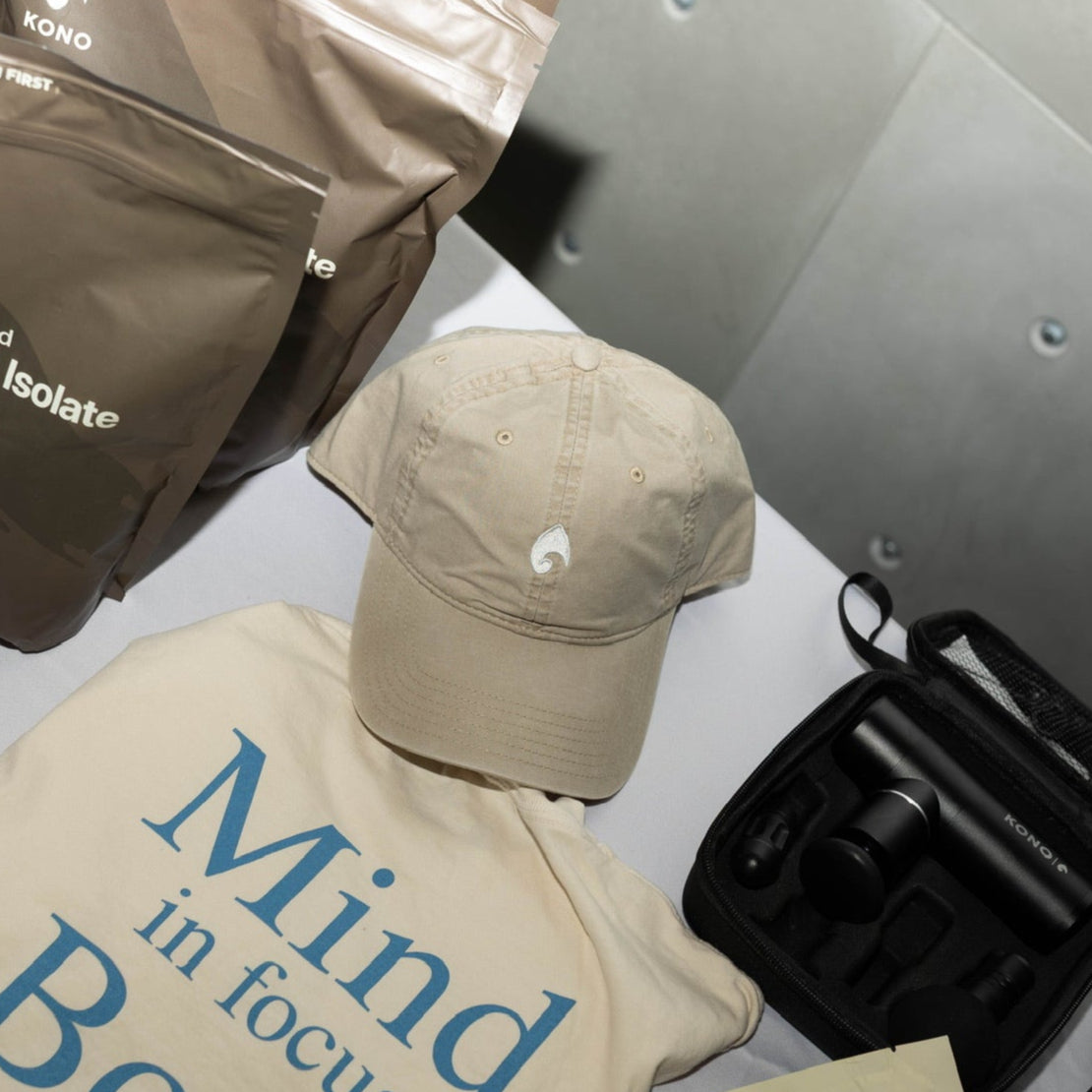 KONO khaki dad hat with embroidered logo styled on a table with KONO whey protein pouches and accessories—lifestyle flat lay.