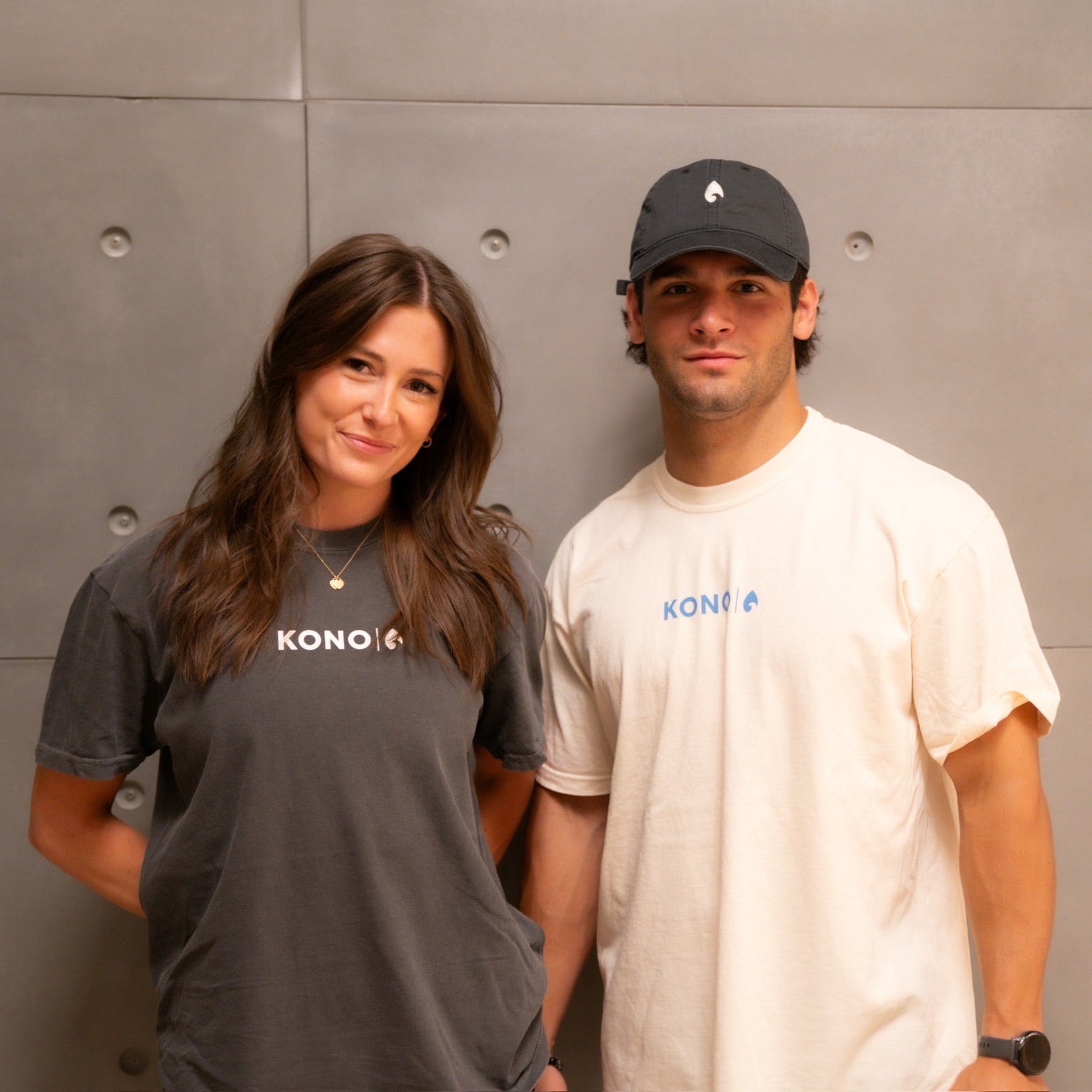 Two models wearing KONO logo t-shirts—charcoal and cream—front graphic, casual fitness apparel against concrete wall.