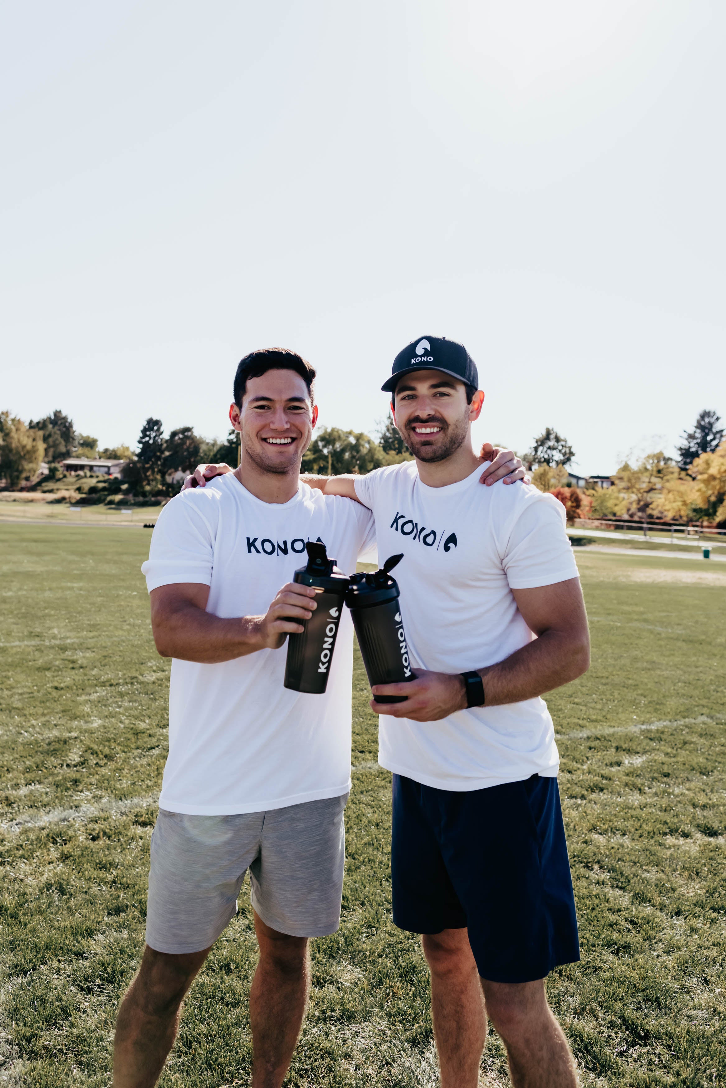 post-workout cheers with KONO shaker cups, two men in white shirts on green turf