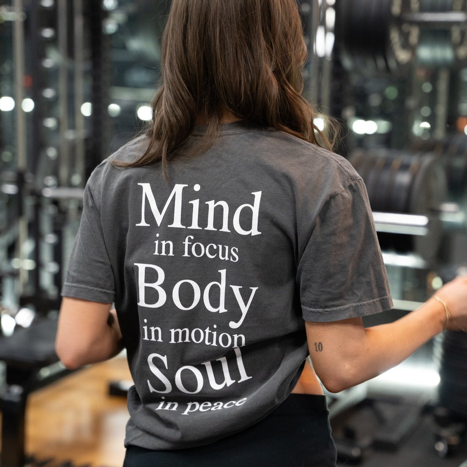 Woman in gym wearing charcoal KONO tee showing large motivational back print—‘Mind in focus, Body in motion, Soul in peace.’