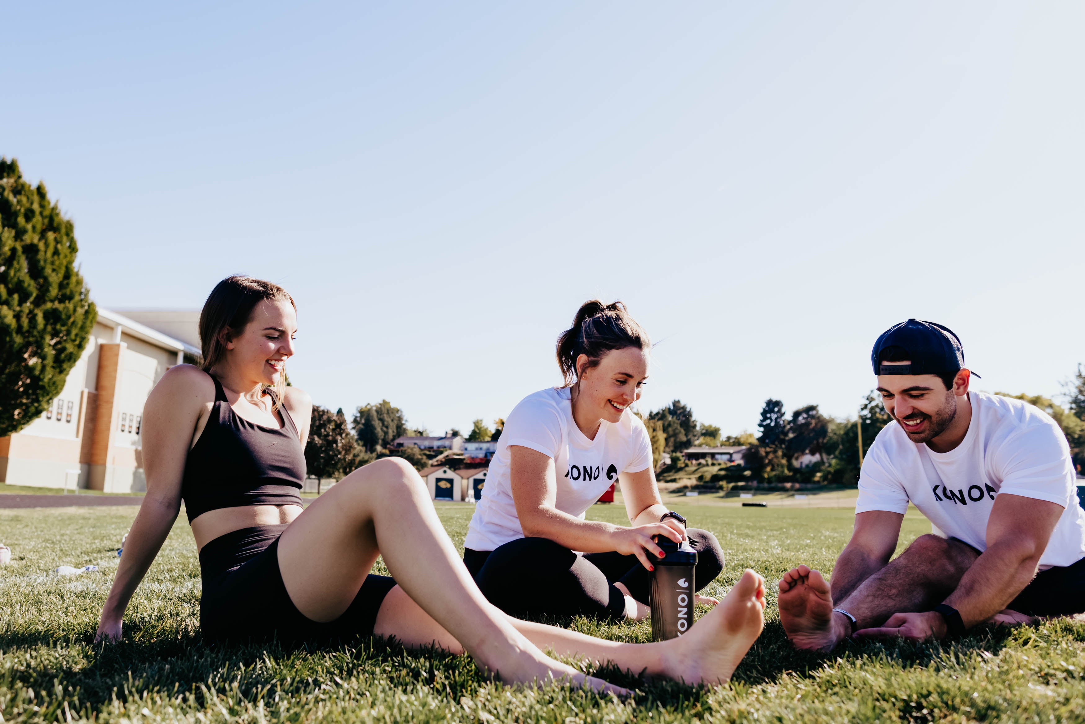 Three athletes sitting on grass stretching and smiling, KONO shaker bottle in the middle, sunny outdoor sports field