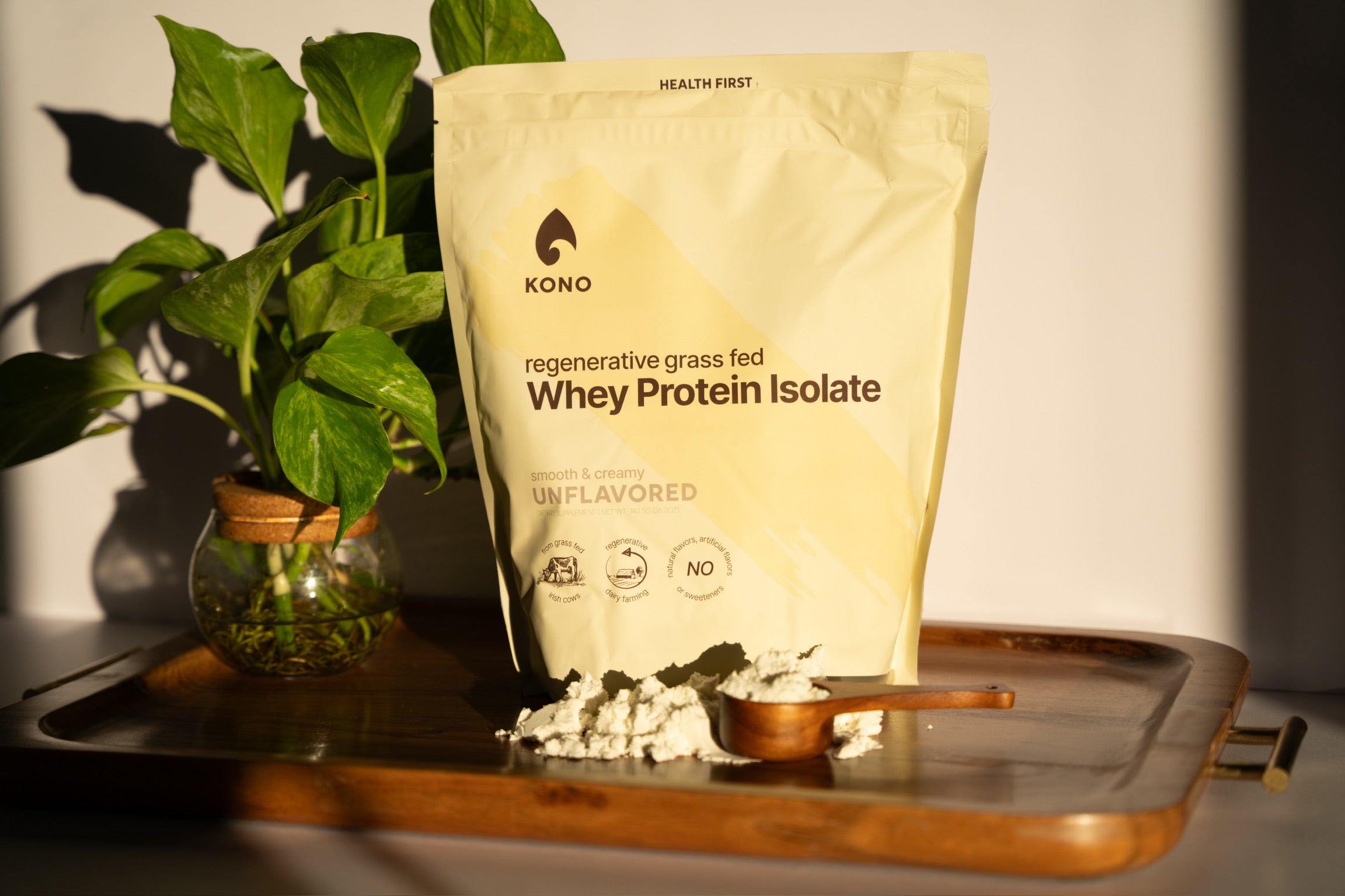 KONO regenerative grass-fed whey protein isolate (unflavored) pouch on a wooden tray with plant and wooden scoop of powder in warm sunlight