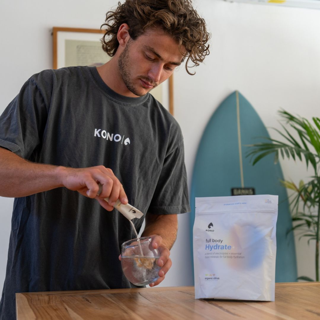 KONO Full Body Hydrate organic citrus electrolyte powder being poured into a glass of water at home, with hydration bag on the counter.
