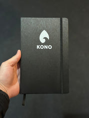 Hand holding a black KONO hardcover notebook with elastic closure and silver logo against a dark studio background