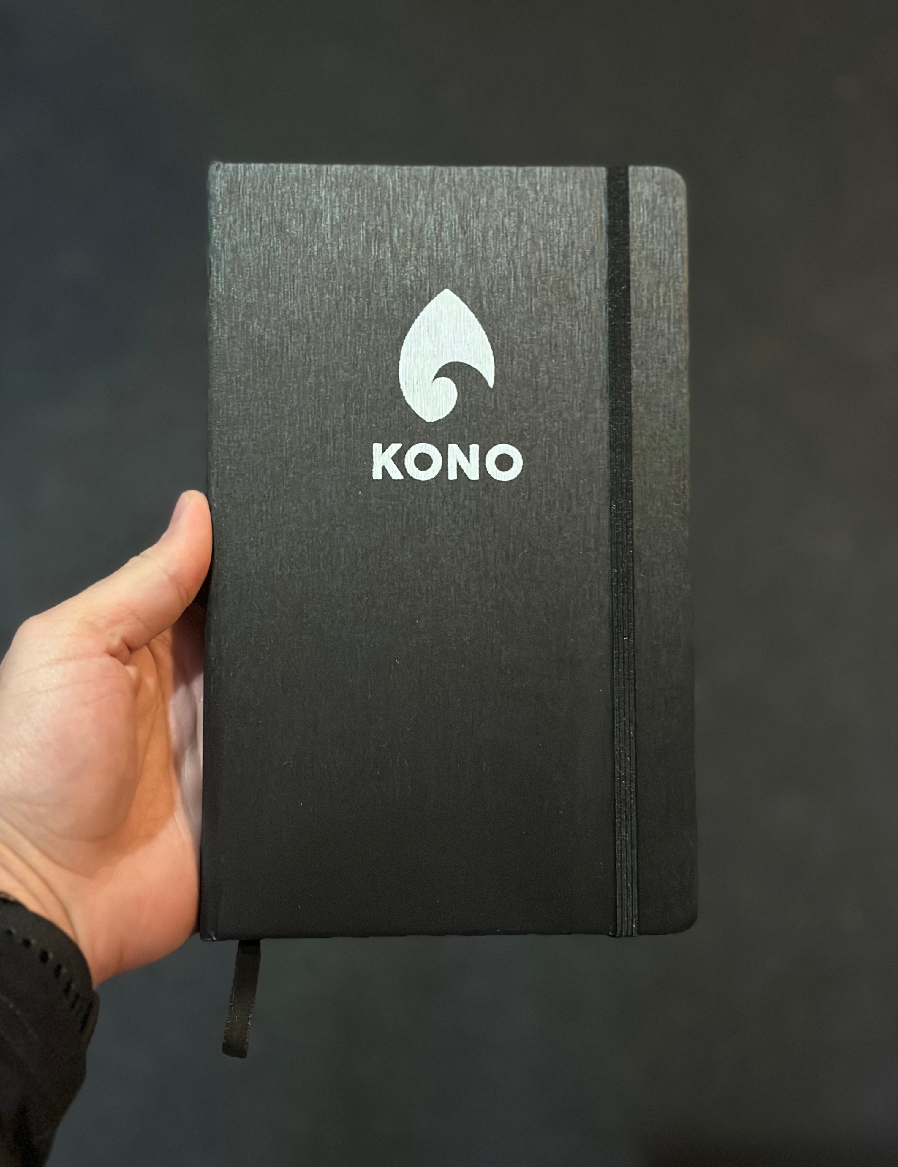 Hand holding a black KONO hardcover notebook with elastic closure and silver logo against a dark studio background