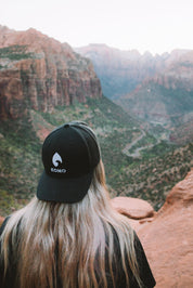 Woman wearing a black KONO embroidered-logo trucker hat overlooking a red-rock canyon—outdoor hiking lifestyle.